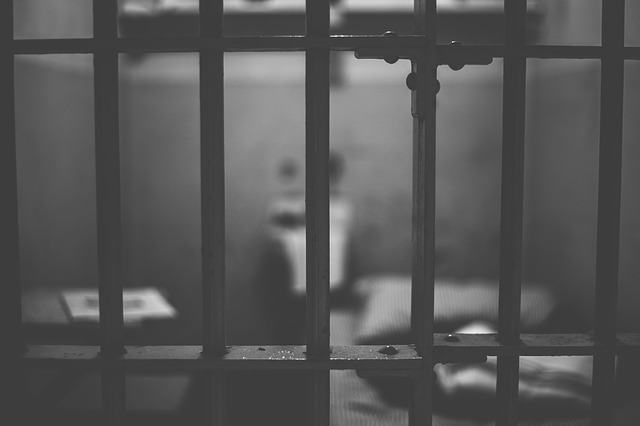 A black and white photo of a jail cell, with the metal bars in focus and everything else out of focus.