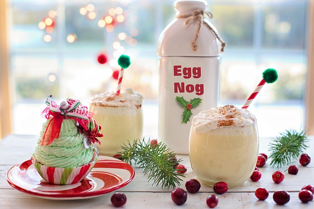 A bottle of egg nog and two cups of egg nog next to a Christmas cupcake and a handful of pine leaves and cranberries
